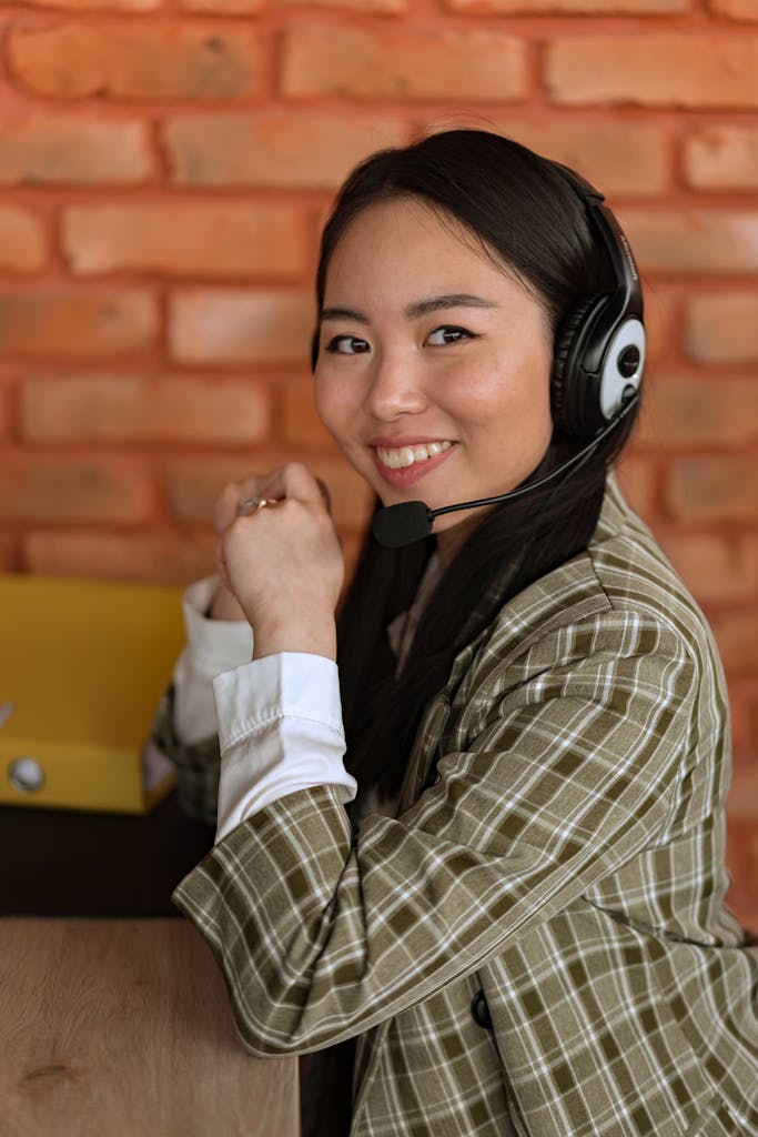 Smiling woman with headset in a plaid blazer, providing customer support in an office with brick wall background.